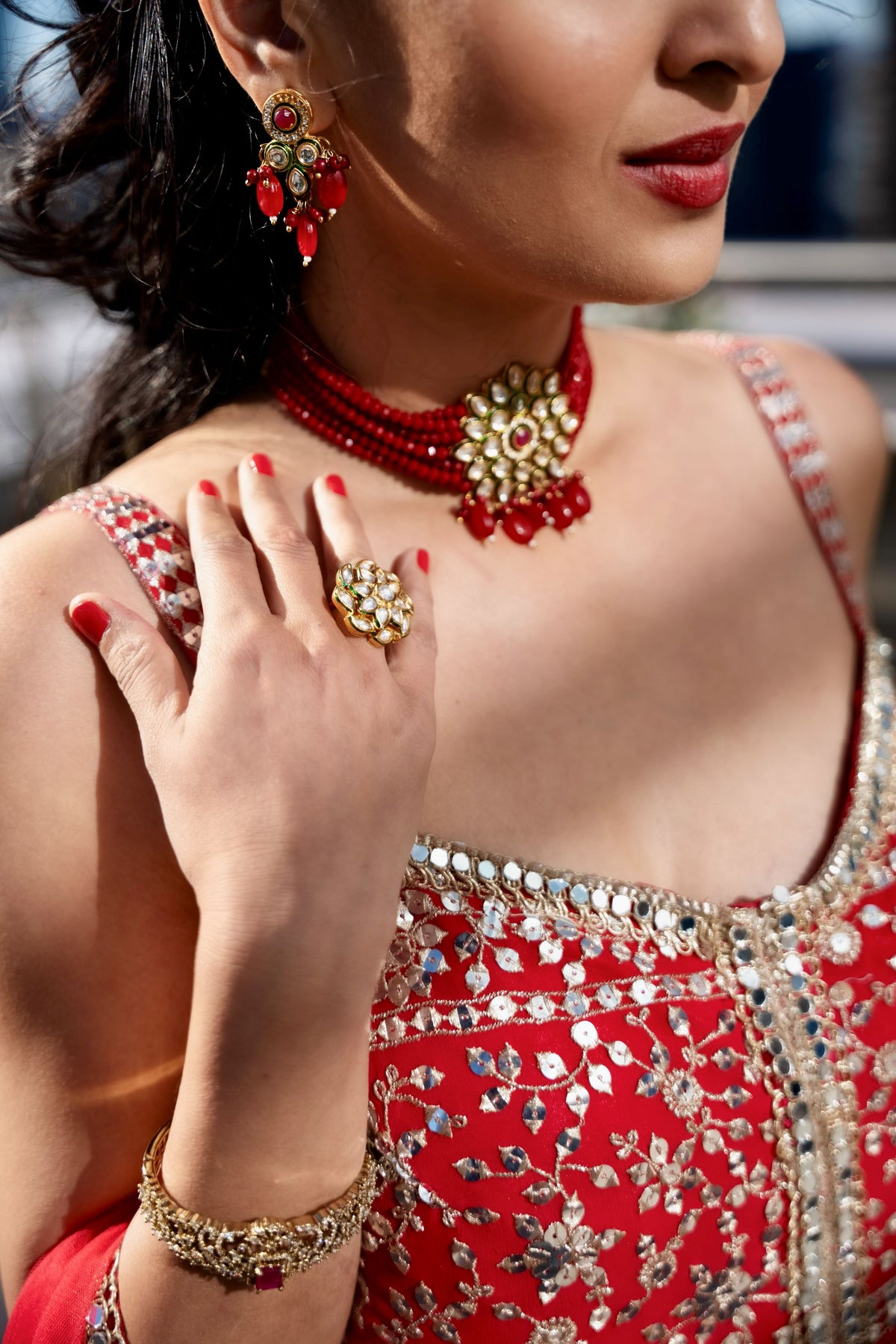 A close-up lifestyle shot of a woman wearing traditional Indian jewelry, featuring a red and gold Posh Pari necklace, matching earrings, a ring, and a bracelet, paired with a red embellished dress.