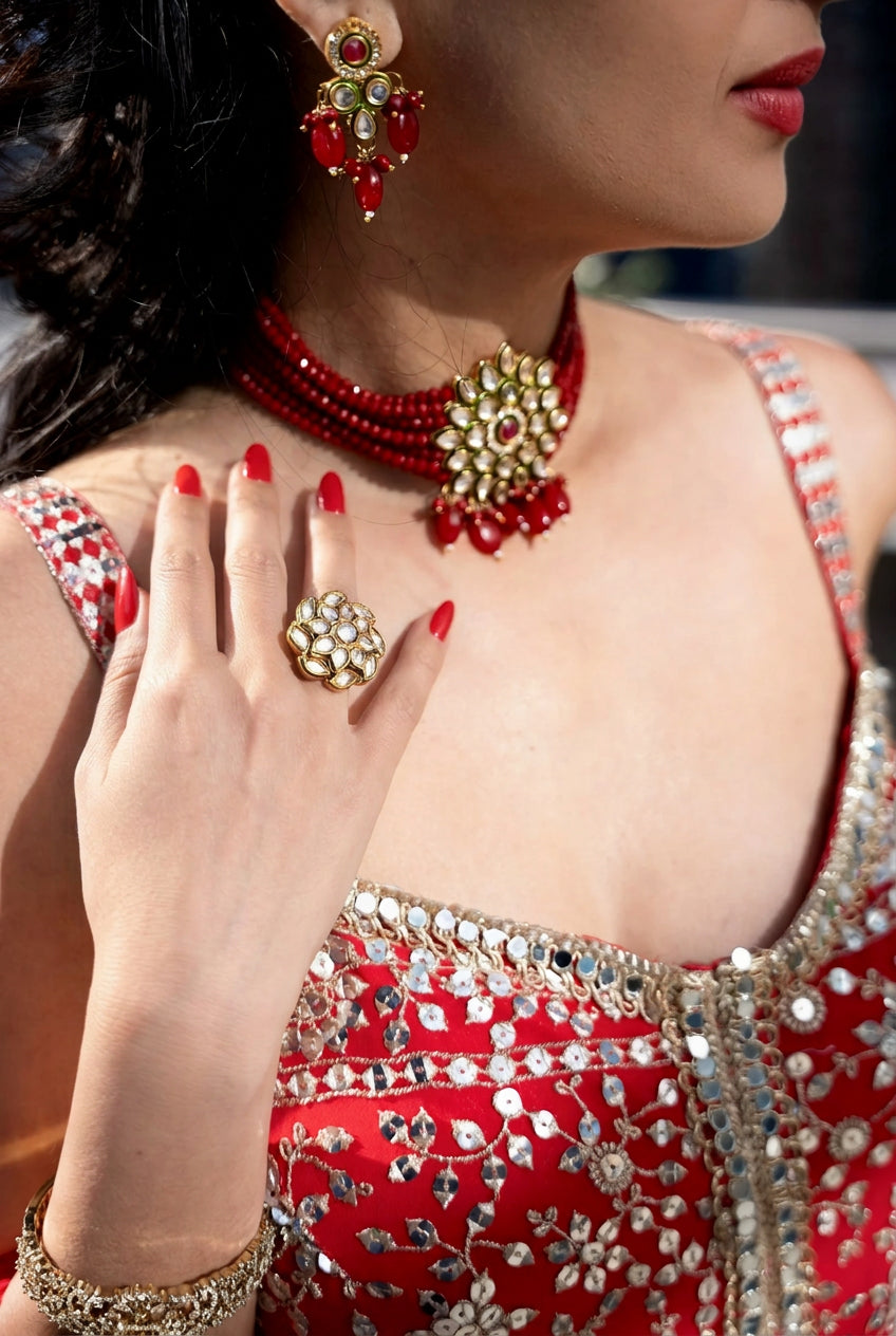 A close-up of a woman wearing a red and gold traditional Indian jewelry set, including a necklace, earrings, and a ring, along with a red embellished dress. all Posh Pari