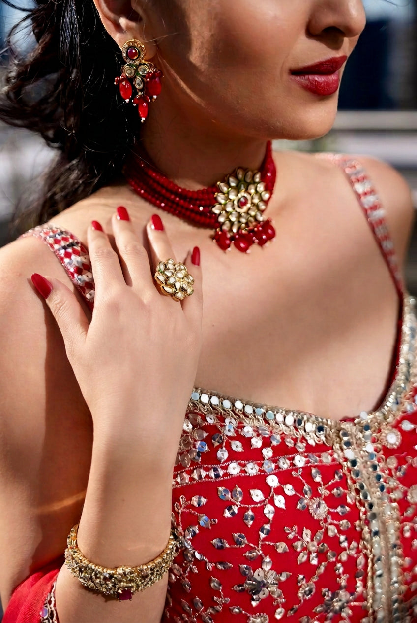 A close-up lifestyle shot of a woman wearing traditional Indian jewelry, featuring a red and gold necklace, matching earrings, a ring, and a bracelet, paired with a red embellished dress. all Posh Pari