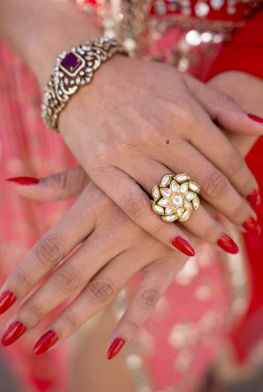A close-up of a person's hands wearing a large gold ring with kundan look stones and a matching ornate gold bracelet, with red nail polish and a red embellished garment in the background. all Posh Pari