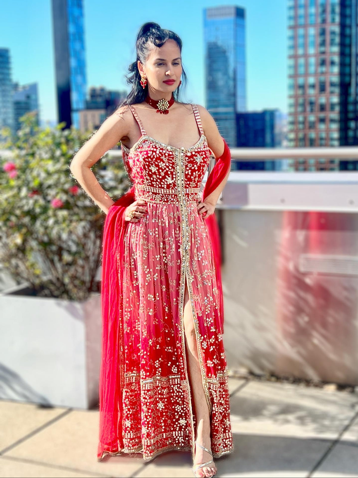 A woman stands on a rooftop terrace wearing a heavily embellished red and pink traditional Posh Pari gown with gold embroidery and a matching red dupatta, with a city skyline in the background.