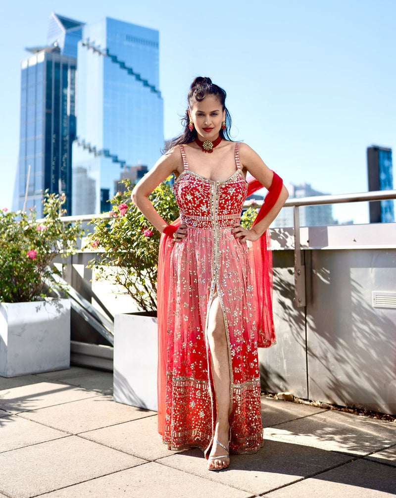 A woman stands outdoors on a rooftop terrace wearing a red and pink heavily embellished traditional Posh Pari gown with a high slit, paired with a matching sheer dupatta. Modern glass skyscrapers and potted plants are visible in the background under a clear blue sky.