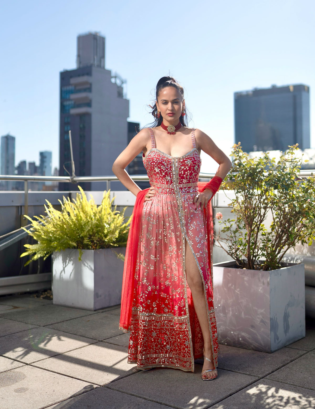 A woman stands on a rooftop terrace wearing a heavily embellished red and gold traditional Posh Pari gown with a high slit, with city buildings and potted plants in the background.
