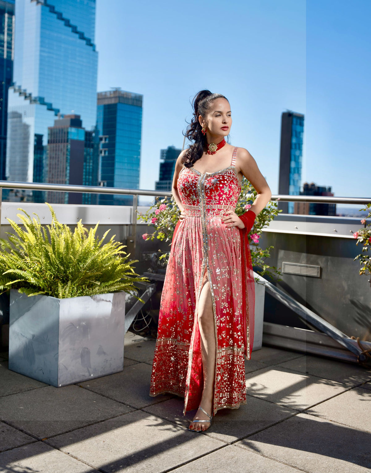 A woman models a red and gold heavily embellished floor-length Posh Pari gown with a high thigh slit, standing on a rooftop terrace with a city skyline and potted plants in the background.