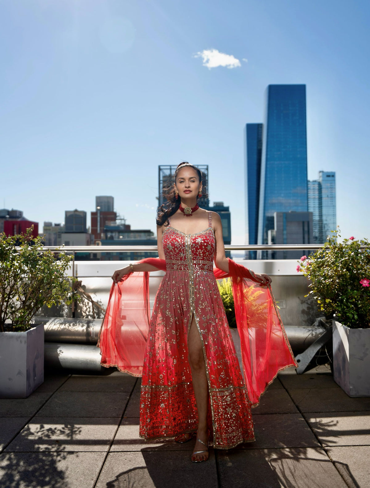A woman stands on a rooftop terrace wearing a sleeveless, heavily embellished red and gold Posh Pari gown with sheer red fabric held out on both sides. The background shows a city skyline with tall blue glass buildings under a clear blue sky.