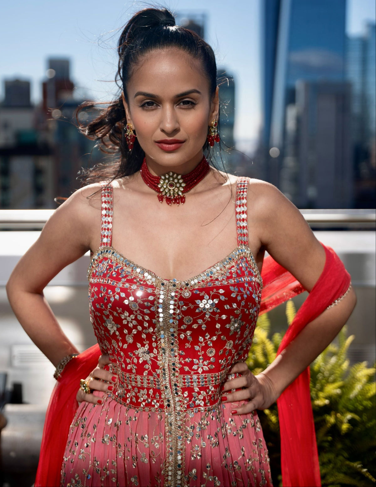 A woman posing outdoors in a heavily embellished red and pink traditional Posh Pari dress with silver embroidery, a red dupatta, and a red Posh Pari choker necklace, with a blurred city skyline in the background.