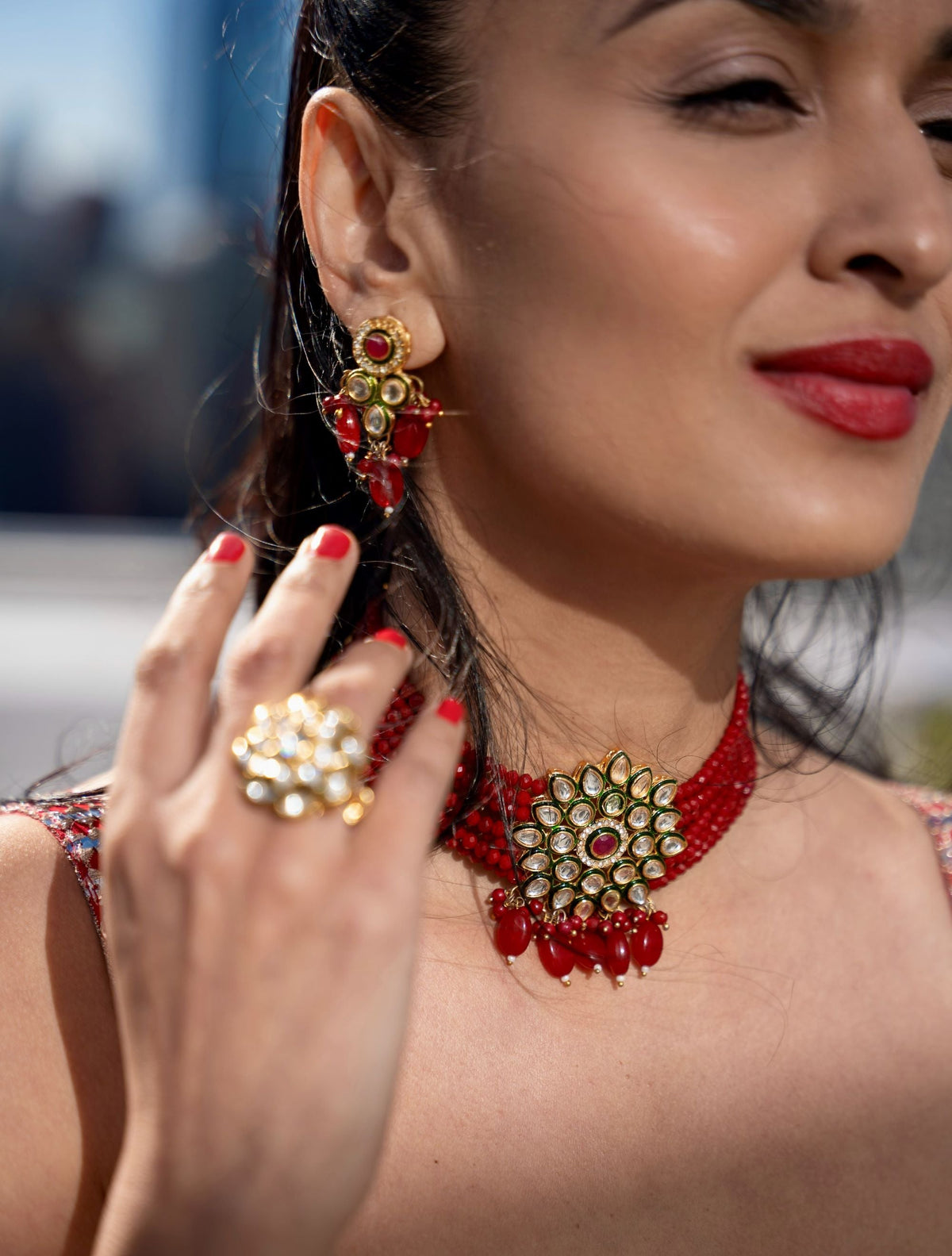 A close-up lifestyle shot of a woman wearing a red and gold traditional Indian Posh Pari jewelry set, featuring a choker necklace, matching earrings, and a statement ring on her finger.