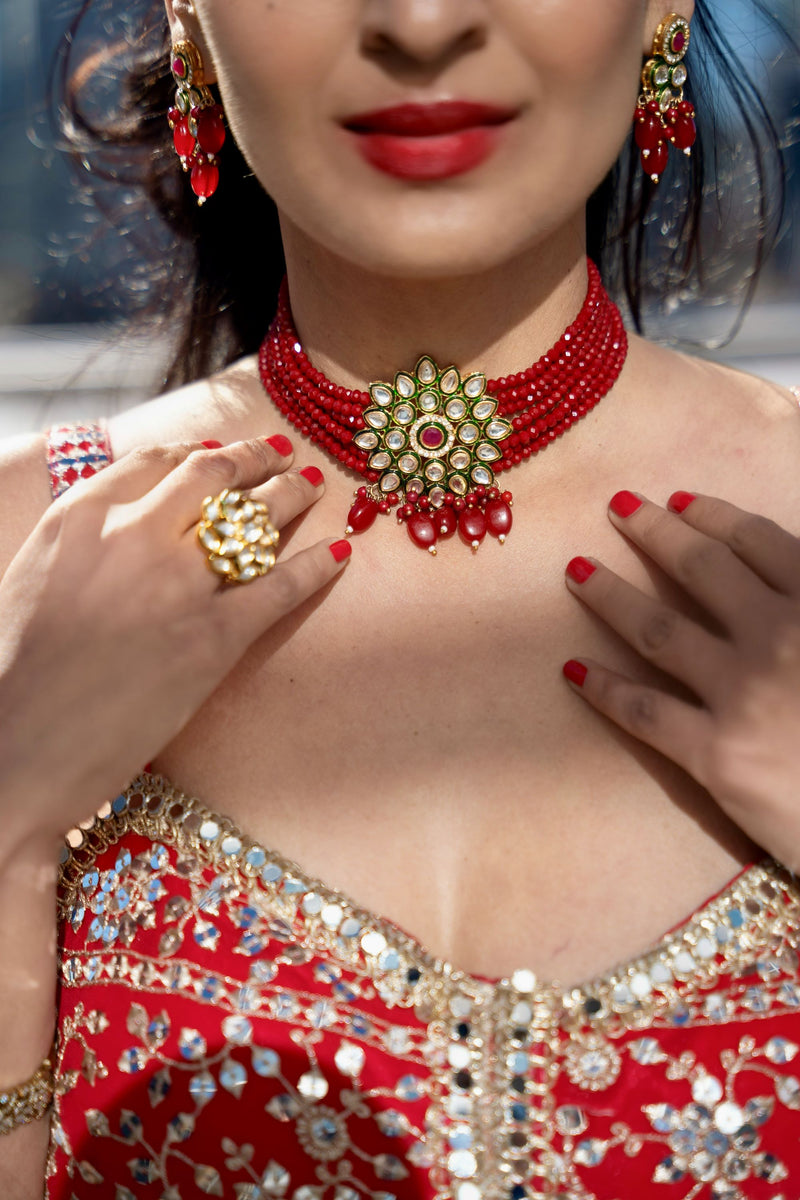 A close-up of a woman wearing a Posh Pari red beaded choker necklace with a large gold-toned floral center and red dangling beads, along with matching red earrings and a red embellished garment.